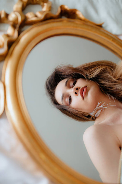 Faszinierende junge Frau mit braunen Haaren und weißen Quasten-Ohrringen, die auf einen ovalen Spiegel mit goldenem Rahmen blickt, der auf hellen Laken liegt. Braut mit nackten Schultern spiegelt sich im Spiegelbild. - Foto, Bild