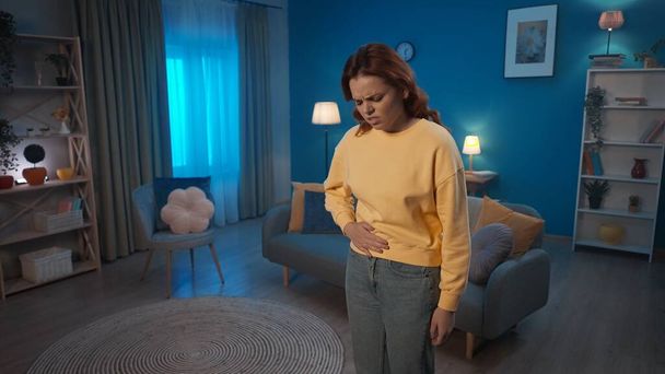 A woman holds her hand on her stomach, suffering from pain, standing in the living room. A woman suffers from abdominal pain, menstrual pain, indigestion, diarrhea. Home medicine concept - Fotografie, Obrázek