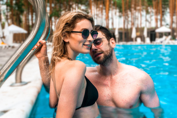 Sorridente donna bionda carina in bikini in piedi insieme a un giovane uomo in una piscina all'aperto. Romanticismo e concetto di vacanza estiva - Foto, immagini