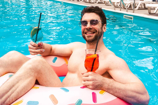 Lächelnder bärtiger Mann mit Sonnenbrille genießt sein Wochenende und hält zwei Cocktails im Swimmingpool. Sommerzeit, Urlaub, Lifestylekonzept. - Foto, Bild