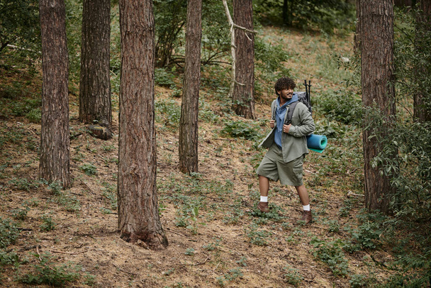 sonriente joven senderista indio con mochila mirando hacia otro lado mientras camina entre árboles en el bosque - Foto, imagen