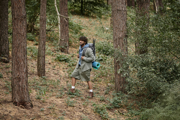 young indian hiker in shirt and shorts holding backpack while walking in forest - Foto, immagini