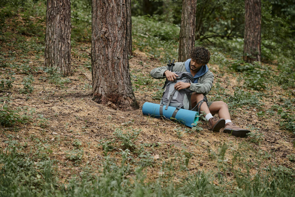lockiger junger indischer Reisender öffnet Rucksack, während er sich ausruht und im Wald auf dem Boden sitzt - Foto, Bild