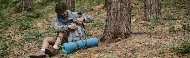 ricci giovane viaggiatore indiano zaino di apertura durante il riposo e seduto a terra nella foresta, banner - Foto, immagini
