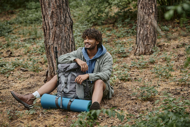 huoleton nuori intialainen turisti lepää istuessaan lähellä reppu maassa metsässä - Valokuva, kuva