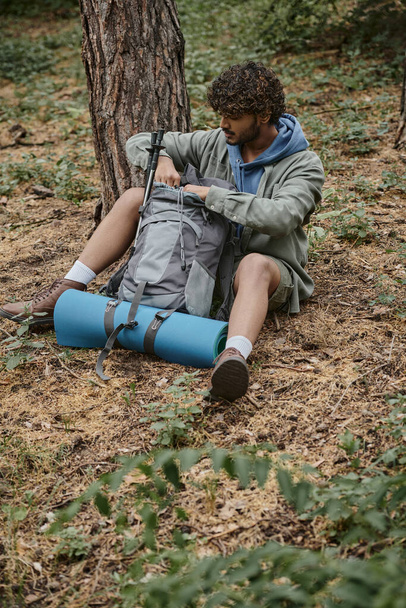 young indian traveler opening backpack with trekking poles and fitness mat in forest - Foto, Imagem