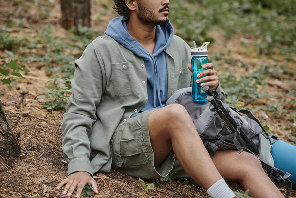 przycięty widok młodego Indianina trzymającego butelkę sportową siedząc w pobliżu plecaka w lesie - Zdjęcie, obraz