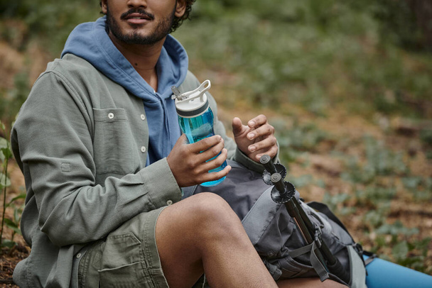 oříznutý pohled na indického cestovatele držícího sportovní láhev v blízkosti batohu v rozmazaném lese - Fotografie, Obrázek