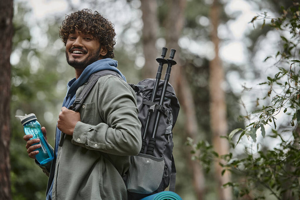 vidám indiai turista hátizsák és sportpalack nézi kamera homályos erdőben - Fotó, kép
