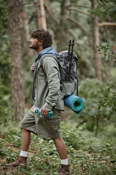 Seitenansicht eines jungen indischen Touristen mit Rucksack und Sportflasche beim Wandern im Wald - Foto, Bild