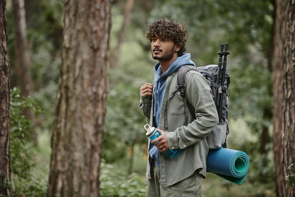 curly young indian tourist in shirt holding backpack and sports bottle in forest - Fotó, kép