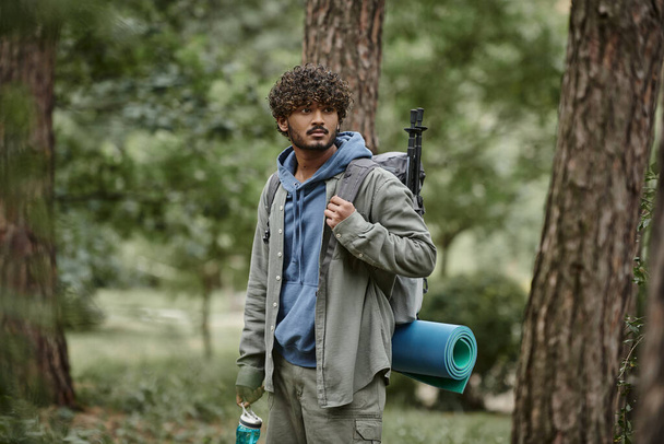 young indian traveler in shirt holding backpack and sports bottle near tees in forest - Photo, Image