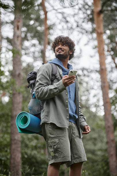 Tiefansicht eines fröhlichen jungen indischen Touristen mit Rucksack im Wald mit Smartphone - Foto, Bild