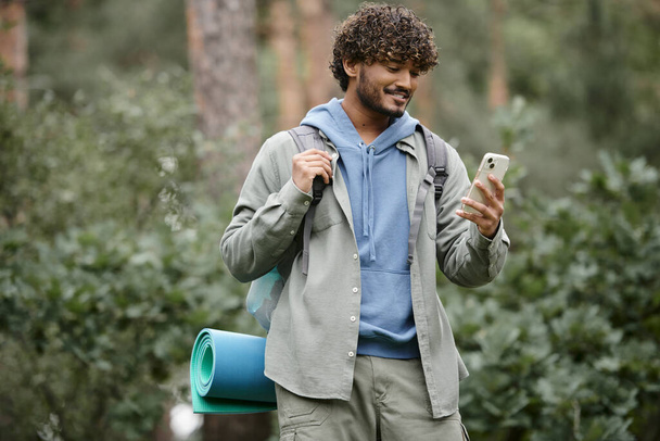 młody uśmiechnięty indyjski turysta z plecakiem za pomocą smartfona podczas stania w zamglonym lesie - Zdjęcie, obraz