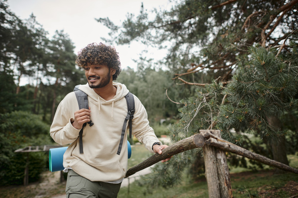 笑顔の若いインドのハイカーとバックパック,ぼやけた森のフェンスの近くを歩く - 写真・画像