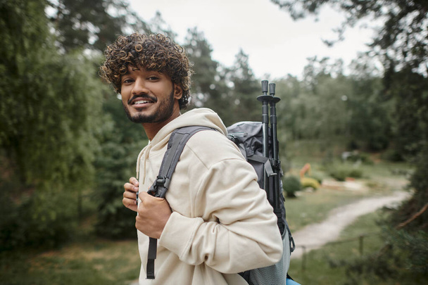 Porträt eines lächelnden jungen indischen Reisenden mit Rucksack, der im verschwommenen Wald in die Kamera blickt - Foto, Bild