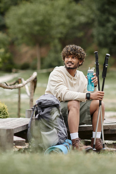 sonriente viajero indio sosteniendo bastones de trekking y botella deportiva cerca de la mochila en el bosque - Foto, Imagen
