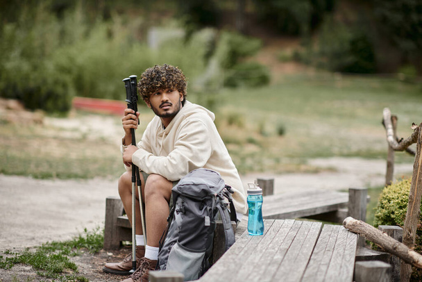 joven turista indio sosteniendo bastones de trekking cerca de la mochila mientras descansa en el bosque - Foto, imagen