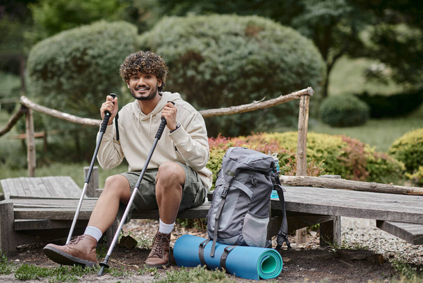 cheerful indian backpacker holding trekking poles and looking at camera in forest - Fotografie, Obrázek