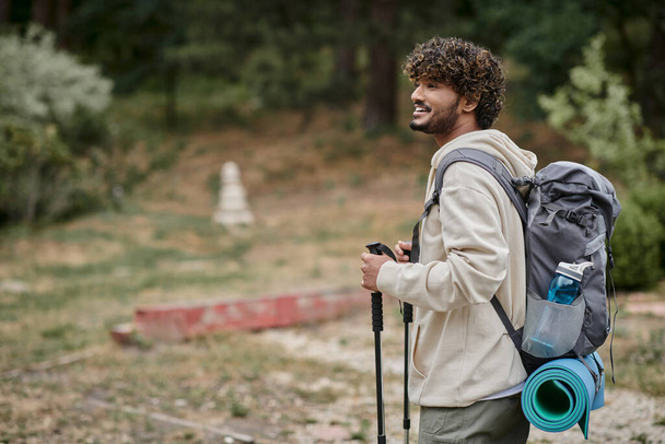 vreugdevolle Indiase backpacker met wandelstokken en weg te kijken in het bos, avontuur concept - Foto, afbeelding