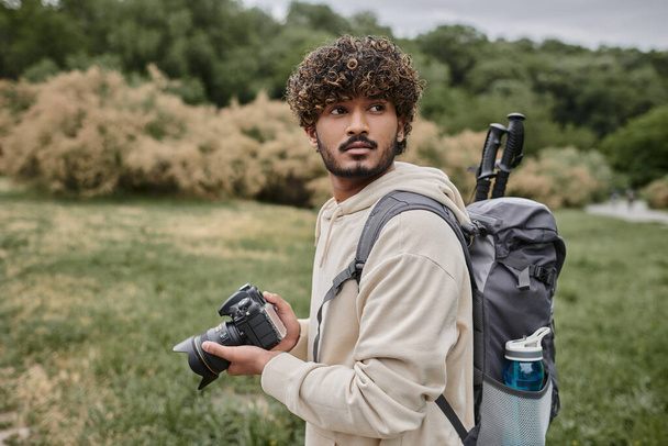 curly indian photographer with backpack holding professional camera outdoors, adventure and travel - 写真・画像