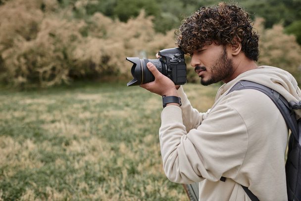 photographe indien concentré avec sac à dos prenant des photos sur appareil photo professionnel, emplacement naturel - Photo, image