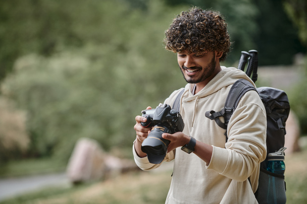 cheerful indian tourist looking at camera and checking photos, hiker with backpack in forest - Photo, Image