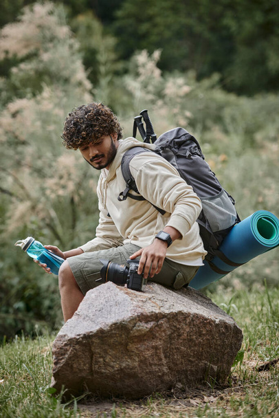 glücklicher indischer Tourist sitzt mit Rucksack auf Felsen und berührender Kamera im Wald, Abenteuer - Foto, Bild