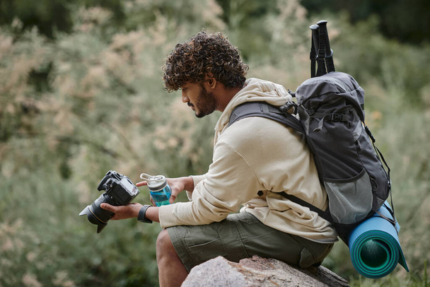 bouclé touriste indien tenant caméra professionnelle et assis sur le rocher, homme avec sac à dos en forêt - Photo, image