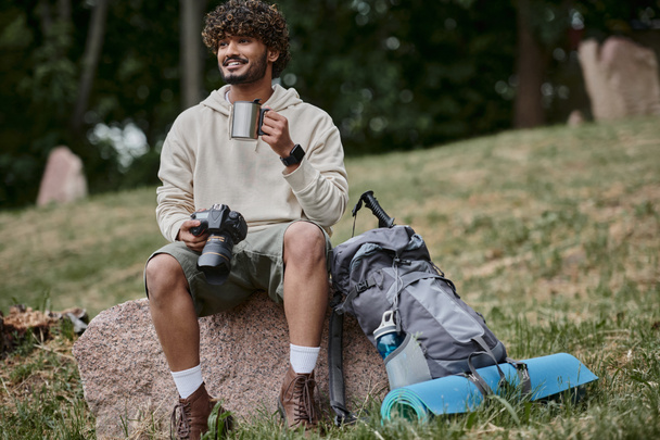 heureux touriste indien tenant caméra professionnelle et assis sur la roche, homme avec thermos tasse dans la forêt - Photo, image