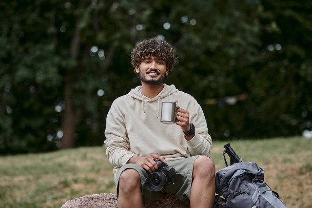 turista indiano segurando câmera profissional e sentado na rocha, homem feliz com copo de garrafa térmica na floresta - Foto, Imagem