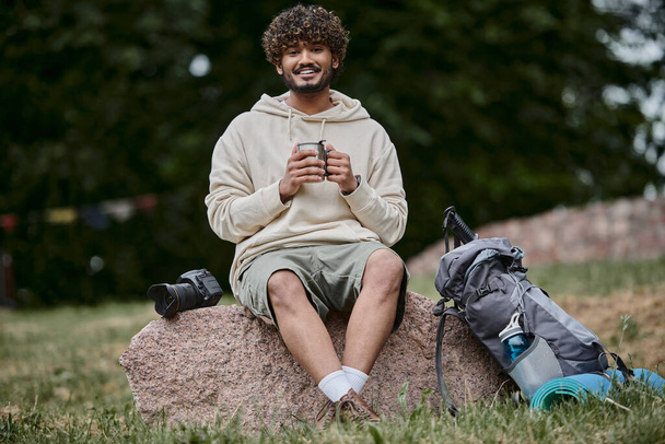 indiai férfi kezében termosz bögre és ül a sziklán, boldog turista néz kamera az erdőben - Fotó, kép
