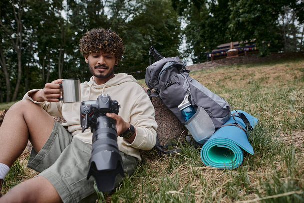 щасливий індійський чоловік тримає термос кухоль і дивиться на фотографії на камеру, турист біля туристичного спорядження - Фото, зображення