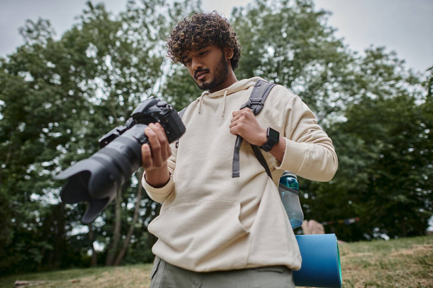 homem indiano positivo segurando câmera digital e de pé com mochila na floresta, fotógrafo - Foto, Imagem