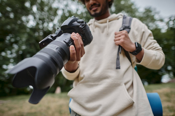 vista recortada del hombre indio sosteniendo la cámara digital y de pie con la mochila en el bosque, fotógrafo - Foto, Imagen