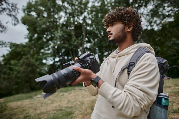 homem indiano concentrado segurando câmera digital e de pé com mochila ao ar livre, fotógrafo - Foto, Imagem