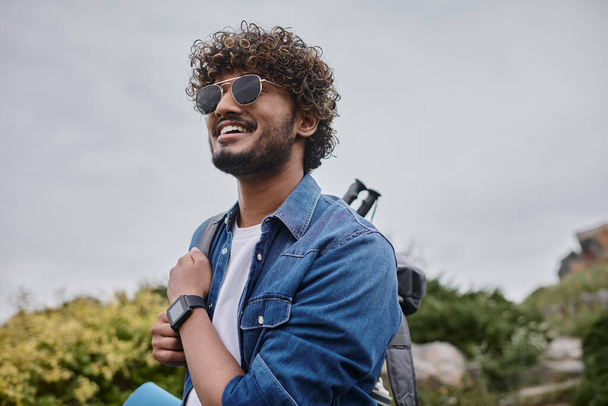 allegro uomo indiano in occhiali da sole in piedi con zaino sulla collina verde, escursionista con attrezzatura da viaggio - Foto, immagini