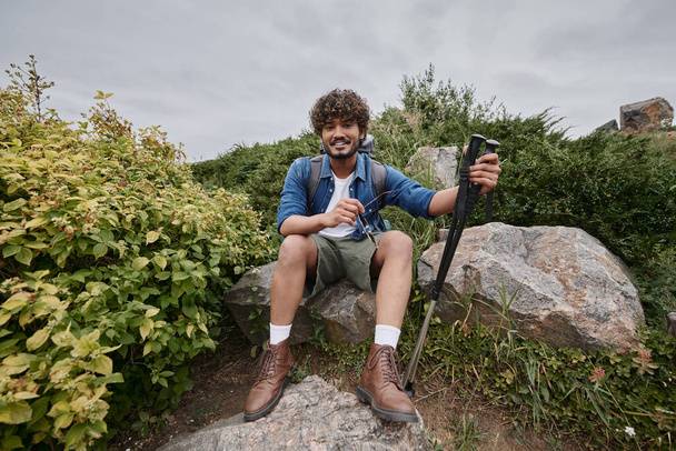 uomo indiano seduto con lo zaino sulla roccia e tenendo bastoni da trekking durante il trekking, avventura - Foto, immagini