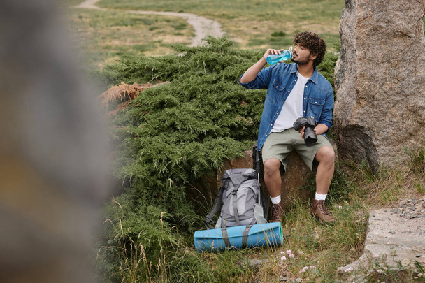 Turist ve doğa konsepti, Hintli adam su içiyor ve kaya üzerinde otururken kamerayı tutuyor. - Fotoğraf, Görsel