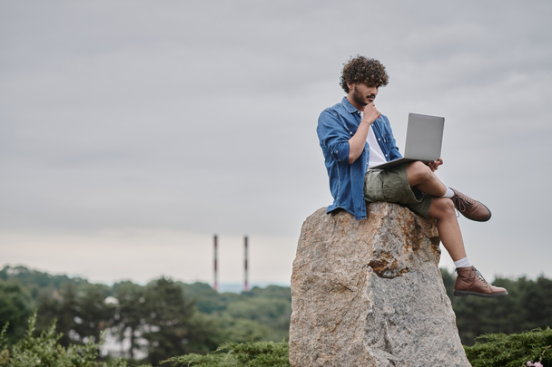 nomade digitale, freelance indiano riccio con computer portatile, seduto sulla roccia, concetto di lavoro a distanza - Foto, immagini