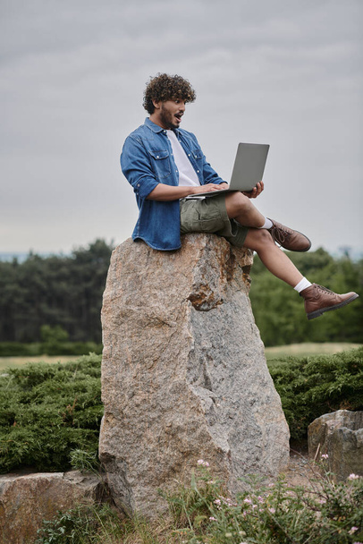 ψηφιακός νομαδισμός, έκπληκτος indian ελεύθερος επαγγελματίας χρησιμοποιώντας το lap-top, που κάθεται στο βράχο, μακρινή έννοια εργασίας - Φωτογραφία, εικόνα