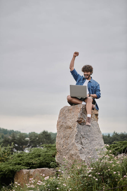 デジタルノマドのコンセプト, 興奮インドの男は,ラップトップを使用しながら勝利を祝い, 岩の上に座って - 写真・画像