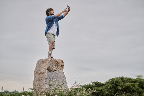 nomadism concept, curly indian man standing on rock and searching signal while holding mobile phone - Photo, Image