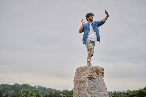 concetto nomadismo, felice uomo indiano in piedi sulla roccia e avendo video chat su smartphone, mano d'onda - Foto, immagini