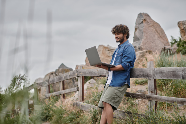 conceito nômade digital, homem indiano alegre em pé perto de cerca de madeira e usando laptop, natureza - Foto, Imagem