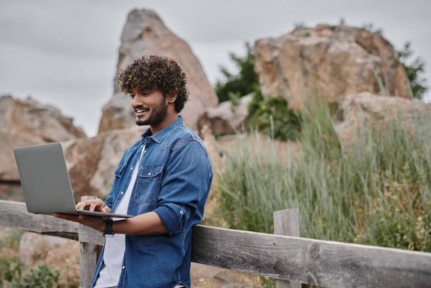 conceito nômade digital, homem indiano alegre em pé perto de cerca de madeira e usando laptop, campo - Foto, Imagem