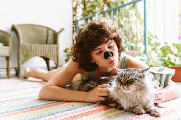 Curly attractive teenage girl is doing yoga, relaxing, sitting on balcony in green top, with beloved cat. Recreation with cat concept - Photo, Image