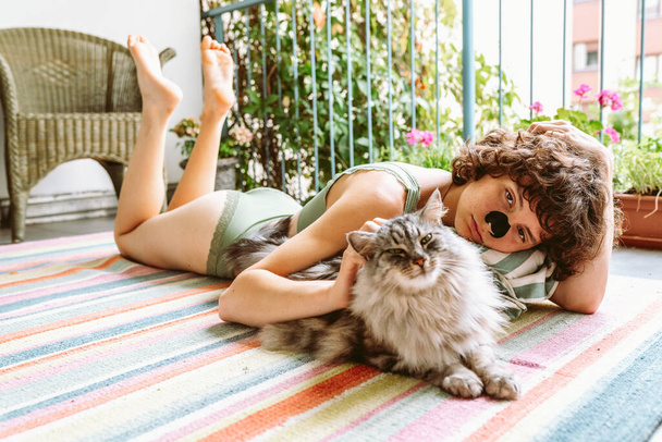 Curly attractive teenage girl is doing yoga, relaxing, sitting on balcony in green top, with beloved cat. Recreation with cat concept - Photo, Image