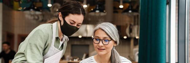 Young waitress woman wearing face mask showing menu to her client in cafe indoors - Photo, Image