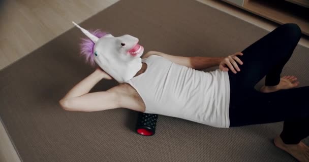 Hombre divertido con máscara de unicornio haciendo ejercicio de espalda con rodillo para la relajación miofascial en casa. Equipo para liberación miofascial y estiramientos humorísticos. Herramientas para auto masaje en casa. - Metraje, vídeo